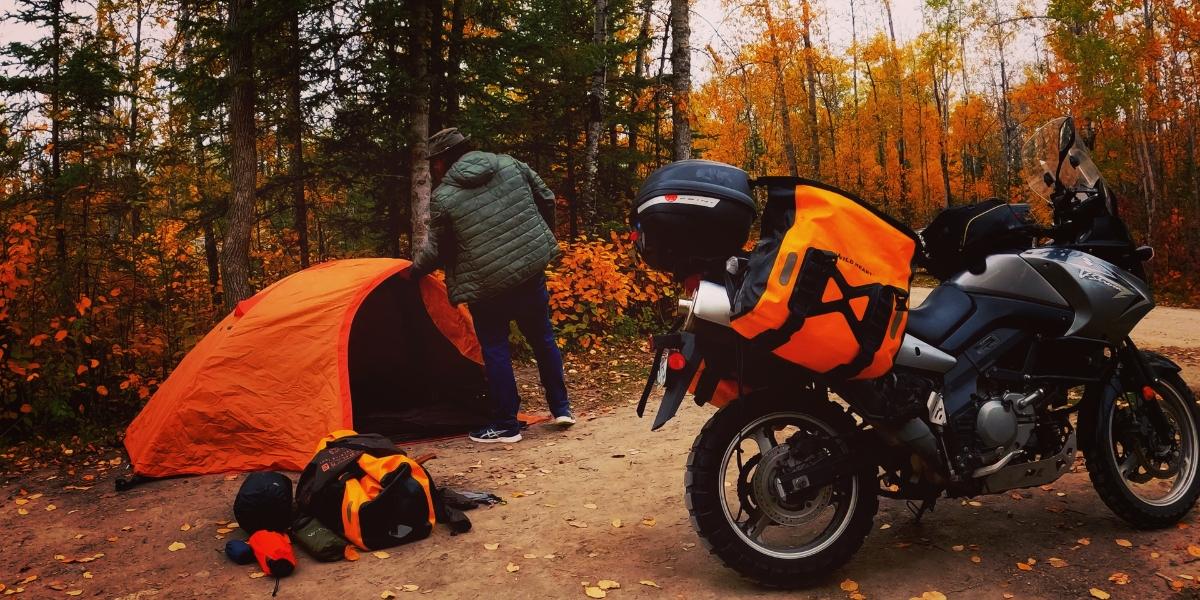 Noel Linsey of Epic Rides MB setting up camp at Nutimik Lake Campground in Manitoba. The image shows Noel pitching his tent with his motorcycle in the foreground.